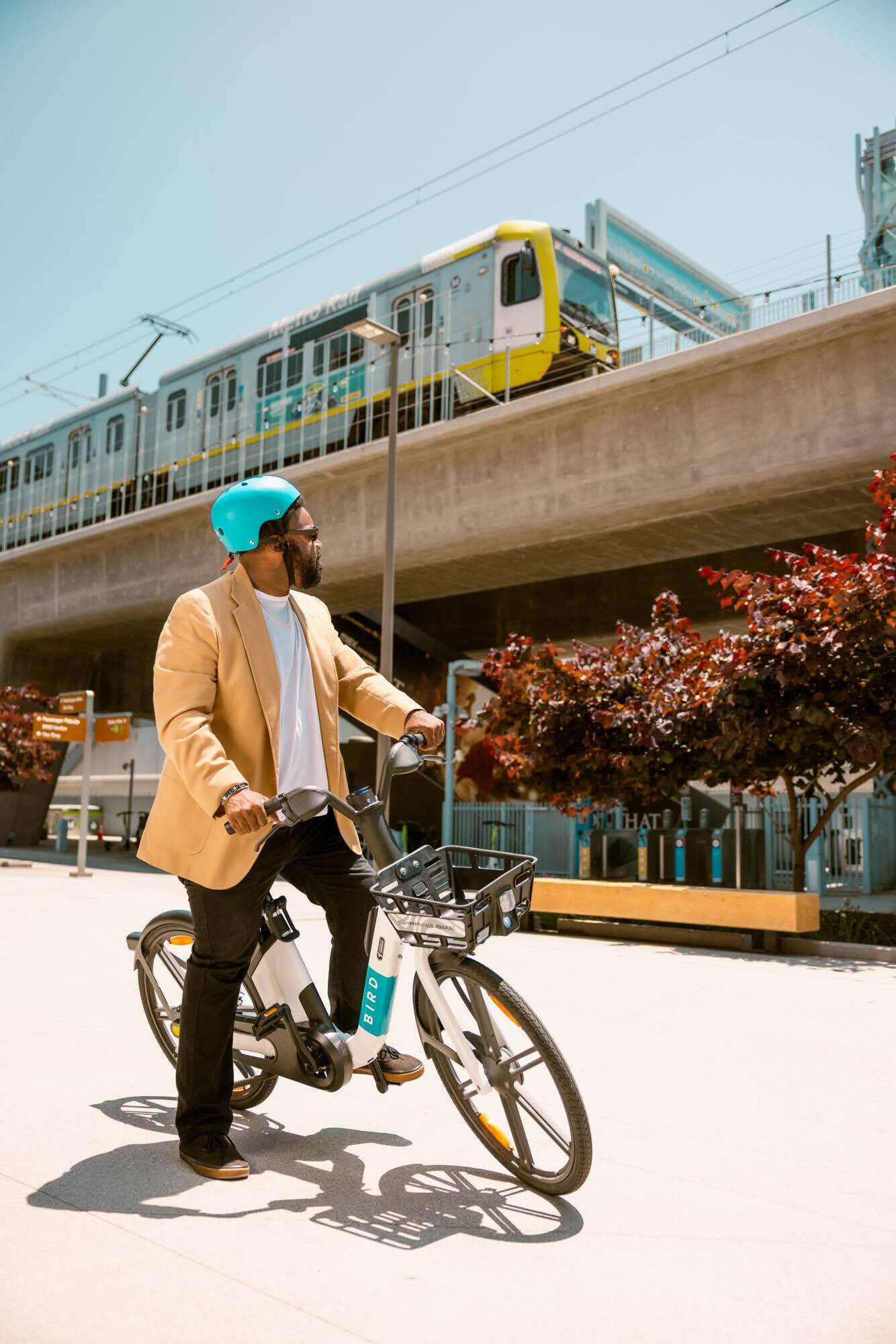 Man in a blazer riding a Bird e-bike with a metro train passing overhead