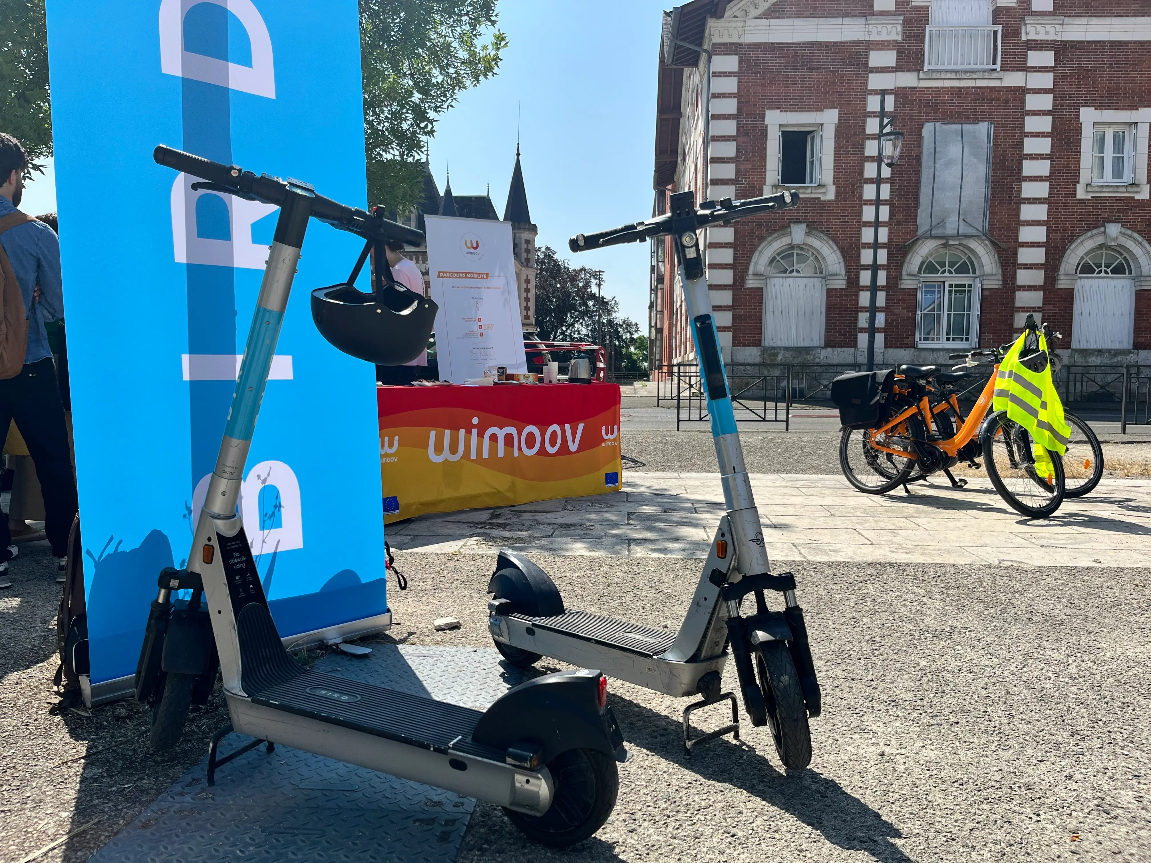 Two Bird scooters parked in front of a Wimoov stand at a mobility awareness event, with Bird bikes in the background.